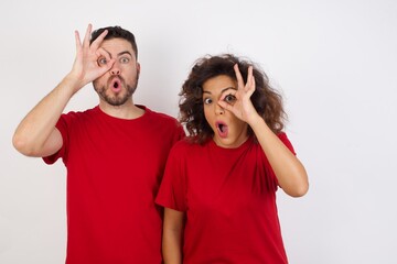 Young beautiful couple wearing red t-shirt on white background doing ok gesture shocked with surprised face, eye looking through fingers. Unbelieving expression.