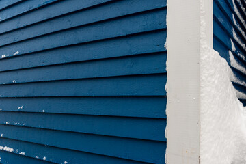 A close up of a bright blue narrow wooden horizontal clapboard on the exterior side of a residential building with a white corner trim board. There's white snow stuck to the textured blue board. 