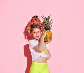 Teenage female with african pigtails, in sunglasses. She is holding pineapple and smiling, posing on pink background. Close up