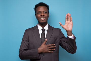 African american man wearing suit smiling swearing with hand on chest and hand up, making a loyalty promise oath. Studio shot on blue wall.