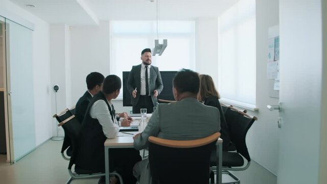 Slow motion tracking shot of stylish bearded man wearing suit standing at table speaking about business plan during meeting in board room