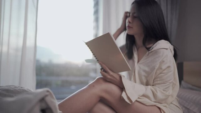 Young Asian lady Sit down reading book alone on the bed, home study turning pages, Reviewing lecture notebook, lesson knowledge, bedroom window day light, self education, Memorize for final exam 