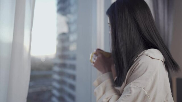 Young Asian woman Having morning coffee after getting up from the bed, female enjoy a cup of coffee and look through the window On high rise condo with urban view, early morning relaxation carefree