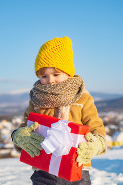 Baby boy in knitted hat holding christmas gift box. Ptoto with empty copy space. Cute little funny child in colorful winter clothes having fun with snow outdoors. Boy in winter mountains.