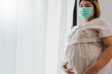 Pregnant Asian women wear a protective mask to prevent germs. Look out of the window During the quarantine at home for preventing coronavirus and covid-19 infection.