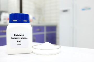 Selective focus of a bottle of BHT or butylated hydroxytoluene chemical antioxidant beside a petri dish with white crystalline powder substance. White laboratory background with copy space.