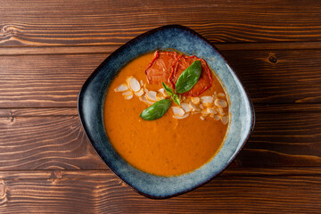 Cold gazpacho with almond leaves and basil on wooden table, top view, blue designer bowl, soft light