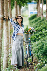Fototapeta na wymiar Happy young Chinese woman standing in park next to her bicyle with basket of groceries in grove or park