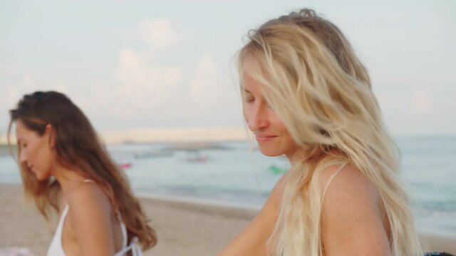 Attractive young woman with long wavy blond hair spending time on beach with her friend waxing surfboard