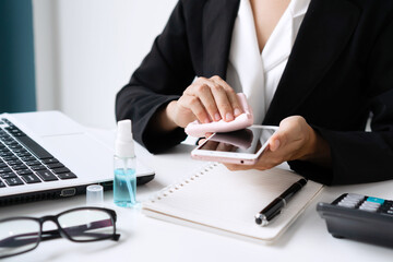Closeup of Asian woman cleaning smartphone by alcohol spray over a work desk in an office. Disinfection daily use item to reduce an infection, preventive measures during the period of pandemic concept