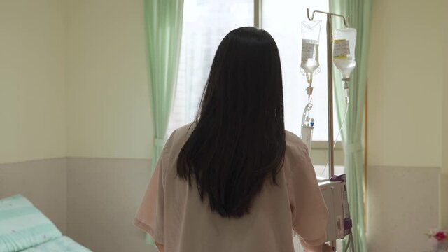 rear view woman holding drip stand is walking slowly to her bed and sitting down. camera movement lonely female patient with iv is returning to her ward