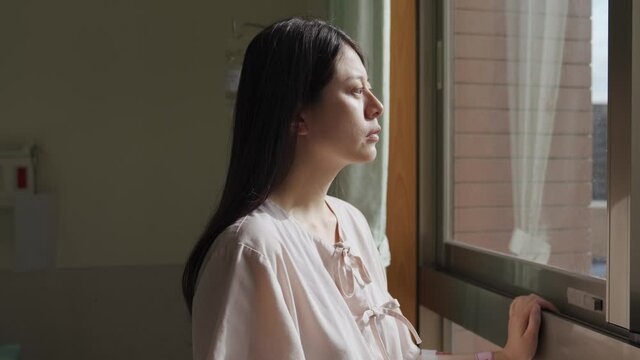 asian woman patient placing hand on window frame looking outside is hoping to get well soon and be discharged