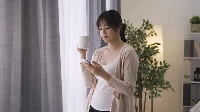 beautiful korean woman sipping coffee by fench window hears notification sound and takes out her cell phone. asian lady is reading and responding phone message from her husband with a smile.
