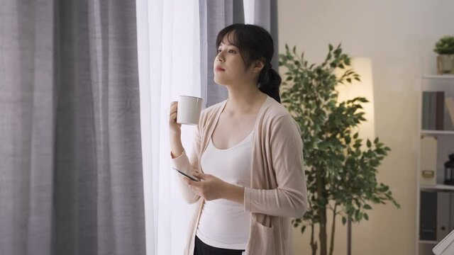 asian young lady relaxing and enjoying tea by curtains receiving message from her besties. elegant chinese female homeowner is texting happily in her spare time.