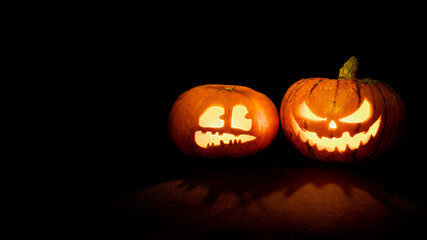Two Halloween pumpkins with glowing eyes and mouth on a black background with copy space. One pumpkin with a scary face, another with a scared face