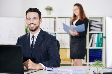 Portrait of cheerful young entrepreneur working on laptop at his office desk, his assistant searching for document in background