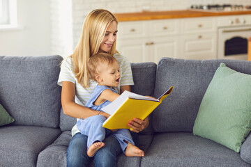 Young mommy reading book of fairy tales to her little child sitting on couch at home