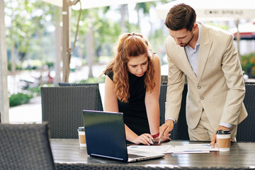 Serious young business people bending over table and discussing details of financial report when...