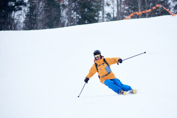 Professional skier concentrated on skiing down on steep ski slope. Proficient technical carving skiing. Net on slope edge. Wooded mountain top on background. Extreme winter activities concept
