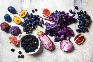 Raw purple vegetables and fruits on gray concrete background. Flat lay of purple food. Eggplant, grapes, figs, plums, blackberries, onions and basil, top view.
