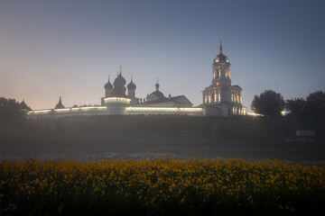 Fototapeta na wymiar A Russian Orthodox monastery on misty sunrise. A meadow with yellow flowers in the foreground. Clear colourful sky. No people