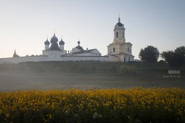 A Russian Orthodox monastery on misty sunrise. A meadow with yellow flowers in the foreground....