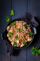 Baked buckwheat with chicken meat and vegetables in black pan, top view, dark background. Traditional Russian cuisine concept.