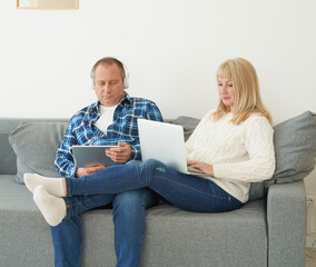 Full length portrait of mature couple with digital addiction at home. Handsome man with tablet and attractive middle age woman with laptop spending time together while lying on sofa. 