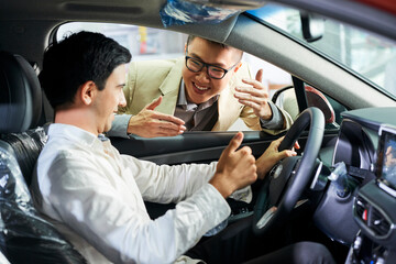 Young man sitting in car salon and showing thumb up while talking with smiling Asian salesman in salon