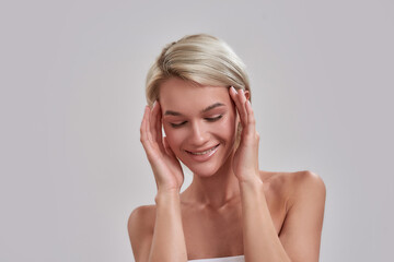 Fototapeta premium Portrait of beautiful young female blonde with perfect fresh skin looking down with a smile, touching her face while posing isolated over grey background