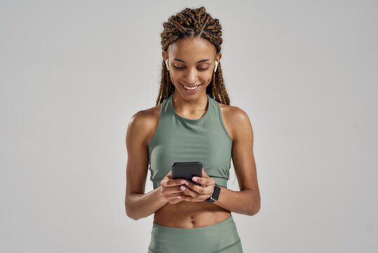 Young positive mixed race fitness woman texting message on smartphone and smiling while standing in studio isolated over grey background, resting after workout