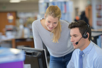lady watching over male colleague wearing headset and using computer