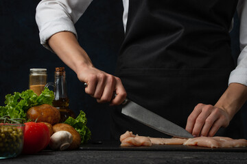 Chef in white shirt and black apron cuts chicken on a dark table isolated on black dark background....