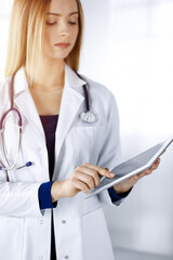 Young professional woman-doctor is using a tablet computer, while standing in a sunny clinic. Portrait of beautiful female physician at work. Perfect medical service in hospital. Medicine concept