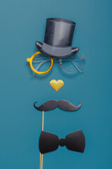 Various black photo booth props: cylinder hat, glasses, moustache, bow tie and nose in heart shape on blue background. Greeting card for father's day. Creative composition in minimal style