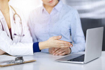 Unknown woman- doctor and her patient are discussing patient's blood test, while sitting together at the desk in the sunny cabinet in a clinic. Female physician, with a stethoscope, is using a laptop