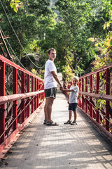 Father son walking across red iron suspension bridge way to forest park. Friendly family vacation outdoor enjoying leisure time nature concept. Dad holds baby by hand, turned around, looking at camera