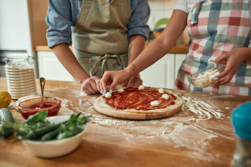 For cheesy taste. Cropped shot of couple making pizza together at home. Man in apron adding, applying tomato sauce on the dough while woman adding mozzarella cheese. Hobby, lifestyle - obrazy, fototapety, plakaty
