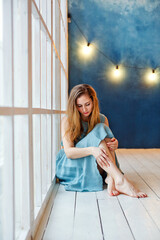 Fototapeta premium A beautiful young woman at the window, sitting on the floor with a sad face, looking away in frustration. Depression