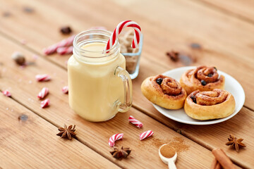 christmas and seasonal drinks concept - eggnog in glass mug with candy cane decoration, cinnamon buns and aromatic spices on wooden background