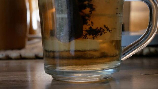 Tea bag inside transparent tea cup just filled with hot water while putting down electric ketle in background
