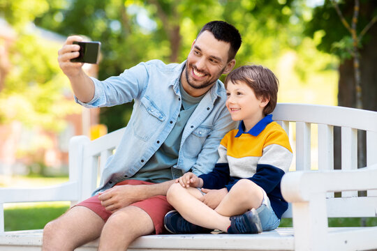 family, fatherhood and people concept - happy father with little son taking selfie picture with smartphone sitting on bench at summer park