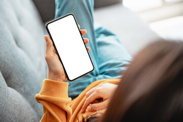 Woman using mobile smartphone with blank white screen on a sofa in living room, copy space.