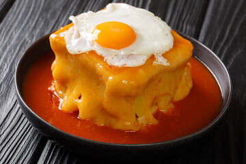 Hot Francesinha sandwich with sausages and meat, melted cheese, poured with tomato beer sauce and topped with a fried egg close-up in a plate on the table. horizontal