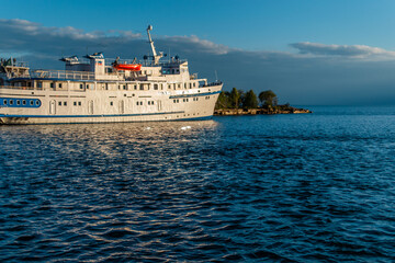 Fototapeta premium white vessel ship boat with blue stripes on pier in bay of lake baikal in light of sunset with reflections in water, trees, horizon