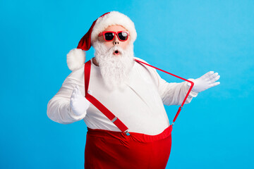 Fototapeta na wymiar Photo of grandpa grey beard hands play straps open mouth surprise wear santa claus x-mas costume suspenders sunglass white shirt headwear gloves isolated blue color background