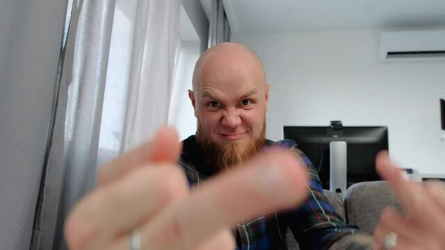 Gestures and signs: Fuck you. A young bald guy with a red beard sitting on the couch shows an indecent non-verbal gesture to the camera. First seriously with one hand, then with both.