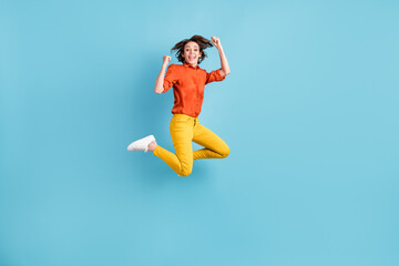 Fototapeta na wymiar Full body photo of attractive funny crazy lady jumping high up fists hands rejoicing best win ecstatic emotions wear orange shirt yellow trousers sneakers isolated blue color background