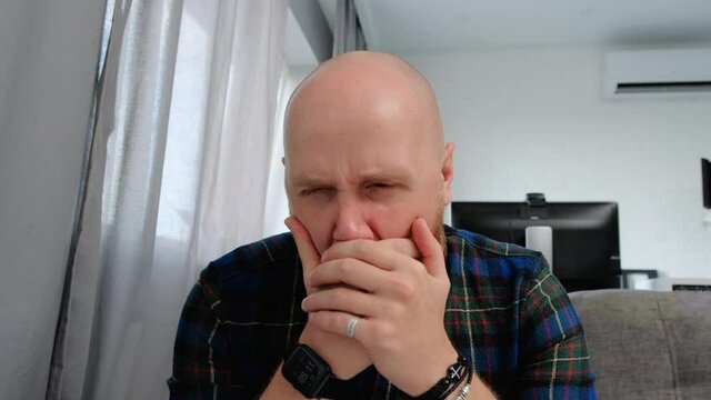 1A young bald guy with a red beard yawns at the camera, covering his mouth with his hand. Close-up of the face. A man sits on a sofa sitting in a modern room.