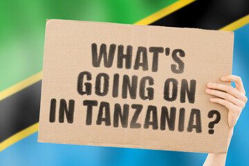 The question " What's going on in Tanzania? " on a banner in men's hand with blurred Tanzanian flag on the background. Protests on the street. Riot. Violence. Economic crisis. Collapse. Politics